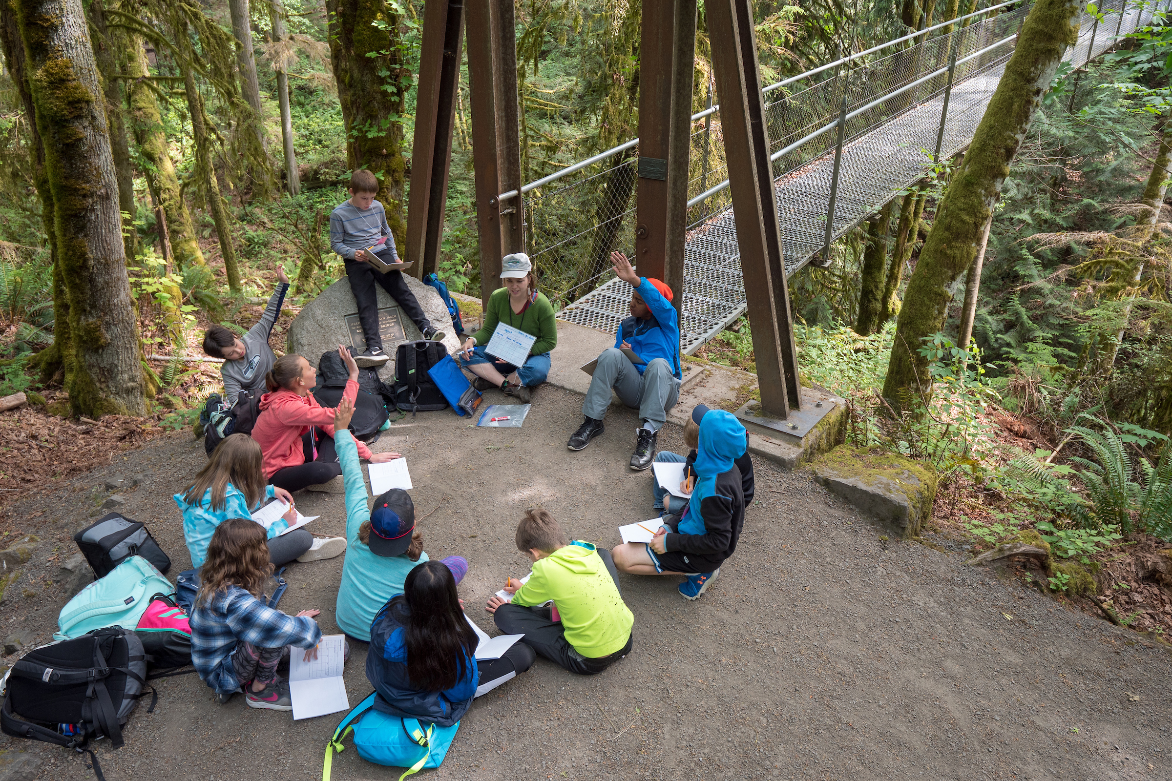 An IslandWood graduate student, in the Graduate Program in Environmental Education & Equity,s sits in a circle with students in the School Overnight Program. Behind them is the suspension bridge