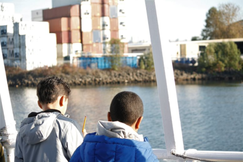 A photo of two students on the Argosy boat during the Duwamish River Program. The backs of their heads are visible and they are looking in the direction of a pile of shipping containers on the bank of the Duwamish River.
