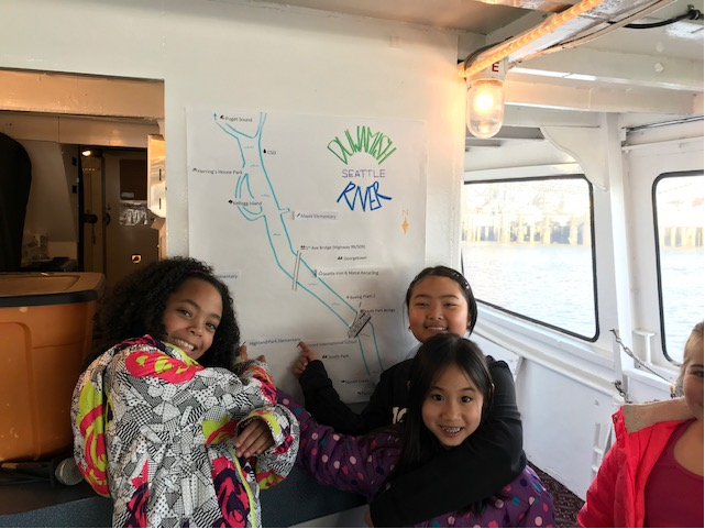 A photo of three students on the Argosy boat during the Duwamish River Program. They are posing in front of a drawn map of the river.