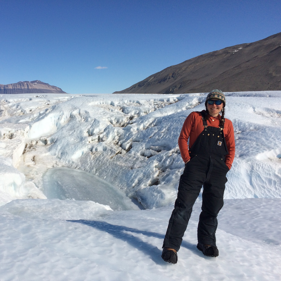 [Image description: Heidi Roop stands with her hands in her pockets on a glacier. She is smiling. Behind her, the sky is blue and hills are visible in the background.]